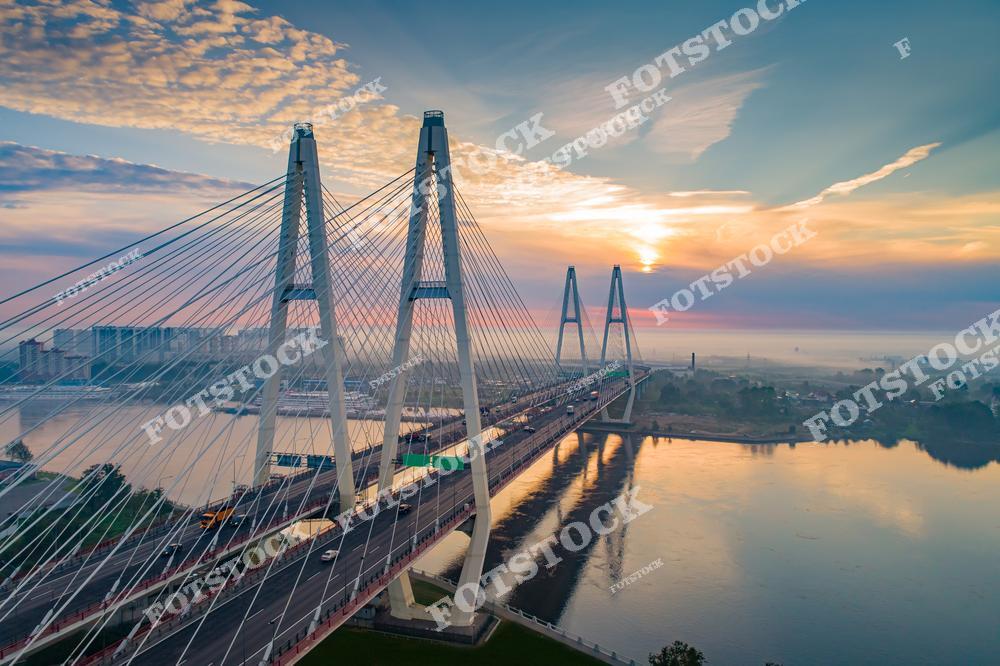 Фотография Санкт-Петербург, Россия, с современной городской архитектурой. На изображении показан вантовый мост, перекинутый через реку Нева, освещённый тёплым светом заката. Фотоснимок передаёт момент вечернего времени суток, когда силуэт моста и его вантовые конструкции эффектно выделяются на фоне неба и воды. Фото отражает инженерное мостостроение России и архитектурный облик Санкт-Петербурга как крупного современного мегаполиса. Иллюстрация может использоваться как фото или визуальный материал, посвящённый мостам и городской архитектуре.
